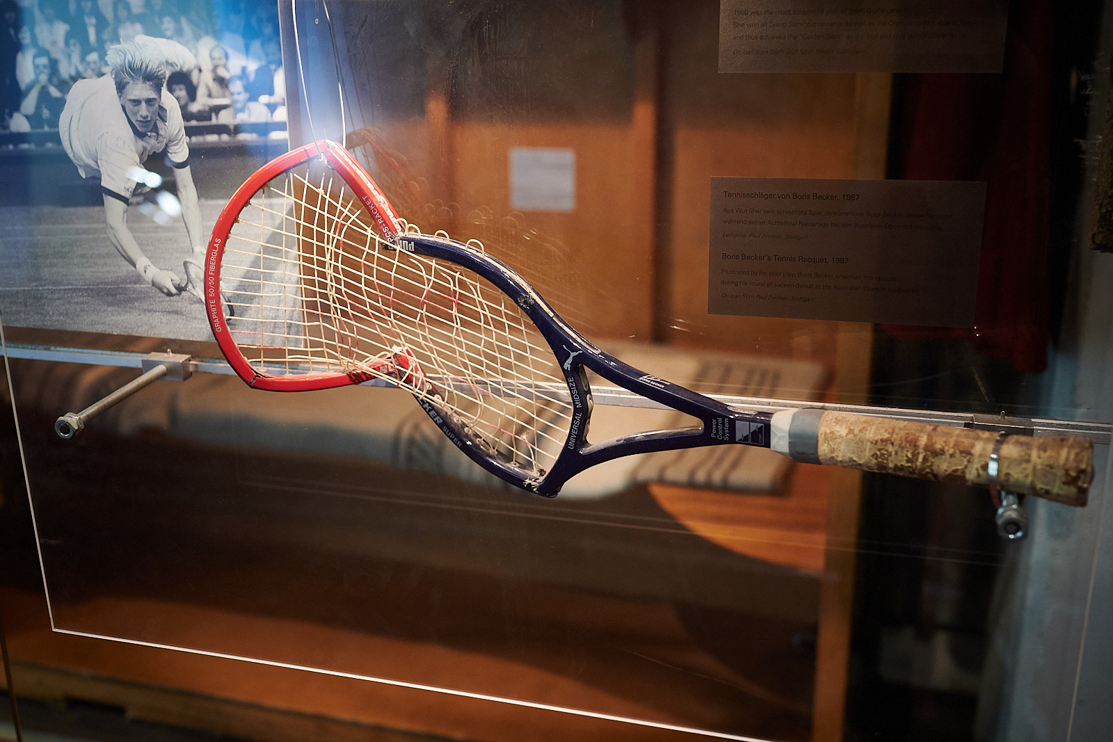 Auf dem Foto sieht man einen Tennisschläger. Es ist der Tennisschläger von Boris Becker. Der Tennisschläger ist kaputt. Neben dem Schläger hängt ein Bild von Boris Becker. Der Schläger wurde an einer Glasscheibe befestigt. Neben dem Schläger ist auch noch ein Bild von Boris Becker.