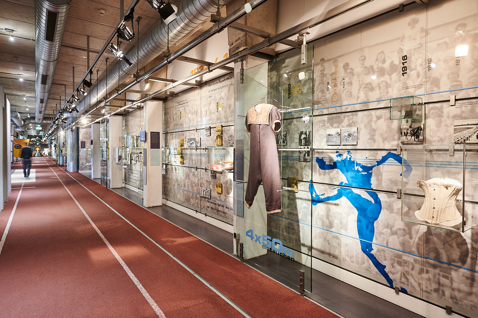 Das Bild wurde in der Dauerausstellung gemacht. Wir sehen einen Teil der Dauerausstellung. Vor uns ist die Zeitleiste von den Olympischen Spielen. An der Zeitleiste sind verschiedene Exponate und Erklärungen. Unter anderem ist auf dem Bild ein alter Badeanzug. Im Hintergrund der Zeitleiste sehen wir eine blaue Figur. Neben der Zeitleiste ist der Boden eine Laufstrecke.