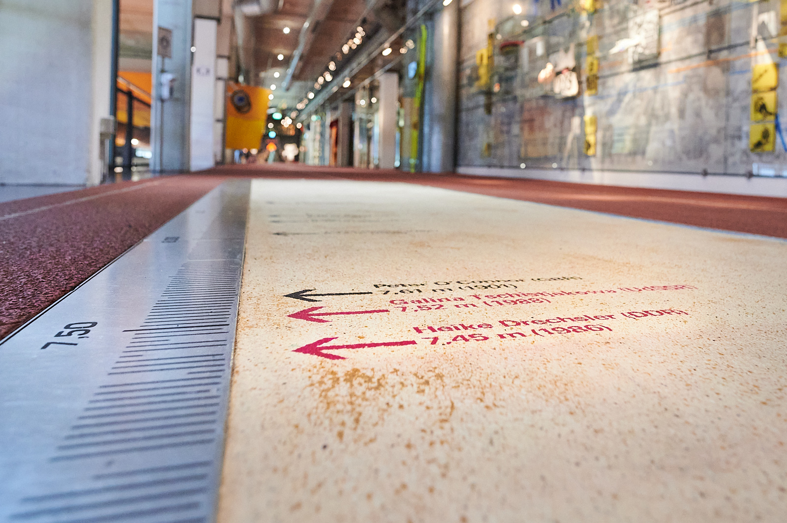 Auf dem Foto sehen wir eine Sandgrube, die aber aus Stein ist. Auf dem Stein sind verschiedene Markierungen. Die Markierungen geben die Weite an, die man gesprungen ist. Auf dem Bild sind drei verschiedene Werte markiert. Im Hintergrund sieht man die Zeitleiste und weitere Teile der Dauerausstellung.