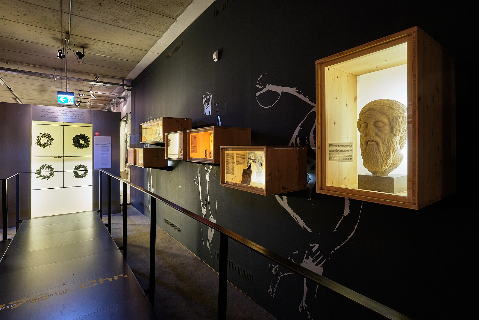 Das Foto stellt den Beginn der Dauerausstellung dar. Es sind verschiedene Ausstellungsstücke aus der Antike zu sehen. An der Wand sind mehrere Kästen angebracht, mit verschiedenen Ausstellungsstücken. Im ersten Kasten sehen wir einen Kopf. Weiter hinten befinden sich verschiedene Kränze.