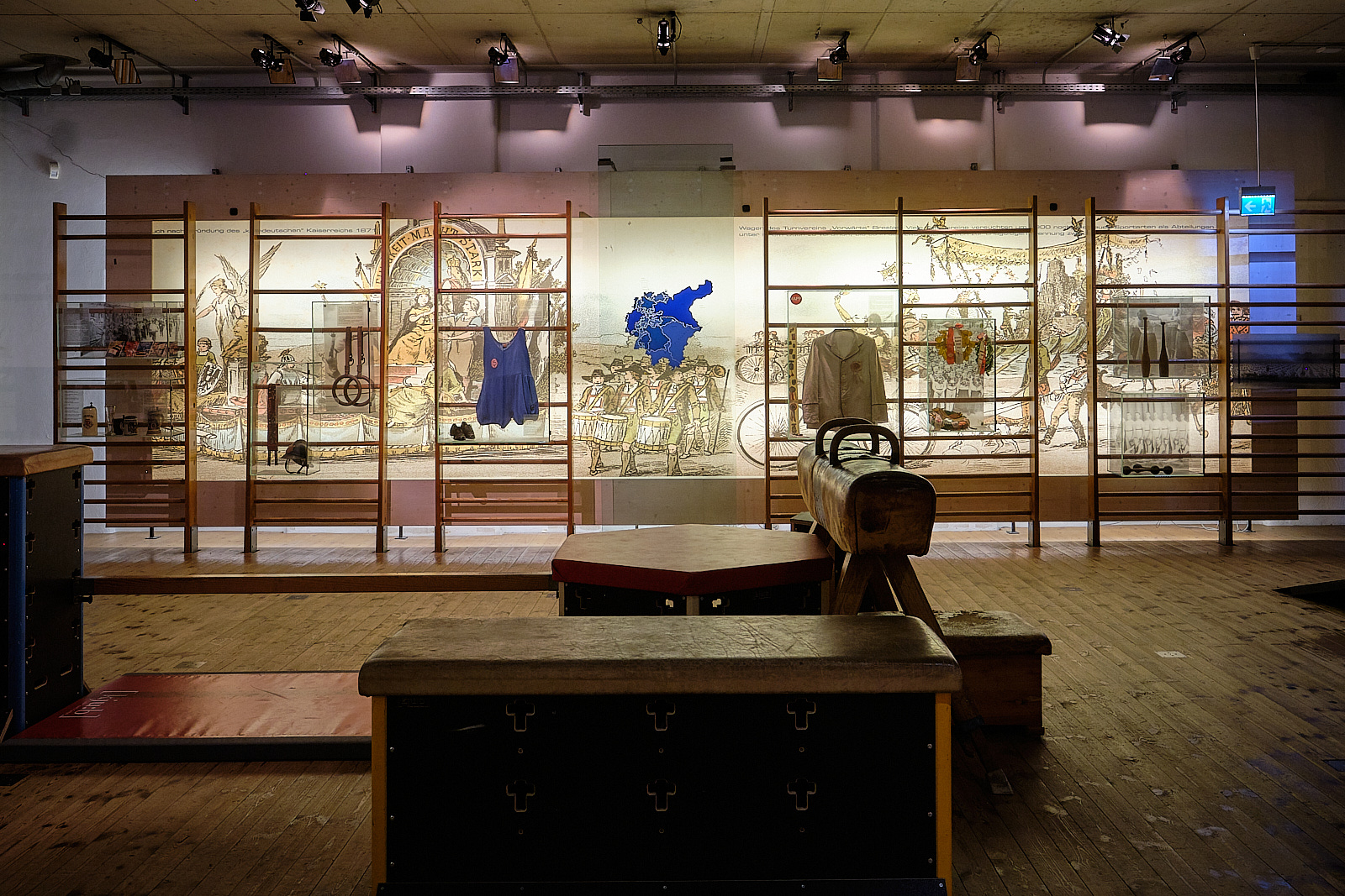 Das Foto wurde in der Turnecke gemacht. Die Turnecke ist dem Turnvater Jahn gewidmet. Man sieht Ausstellungsstücke an der Wand und verschiedene Sprossenwände. Es sind nur wenige Lichter an. Dafür ist die Wand beleuchtet. Außerdem befinden sich verschiedene Turngeräte im Raum.