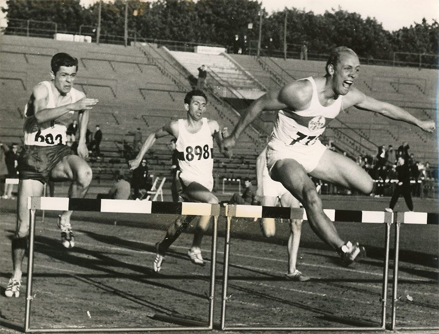 Fotografie in Schwarz und Weiß von Willi Holdorf im Sprung über eine Hürde. Im Hintergrund zwei weitere Hürdenläufer