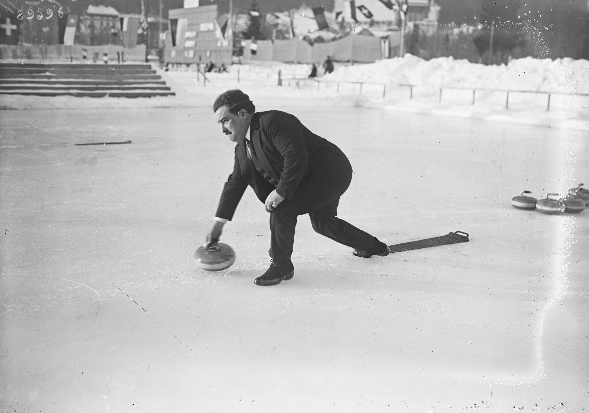 Ein Mann ist zu sehen, der einen Curling-Stein über das Eis schiebt. Er ist im schicken Anzug gekleidet.
