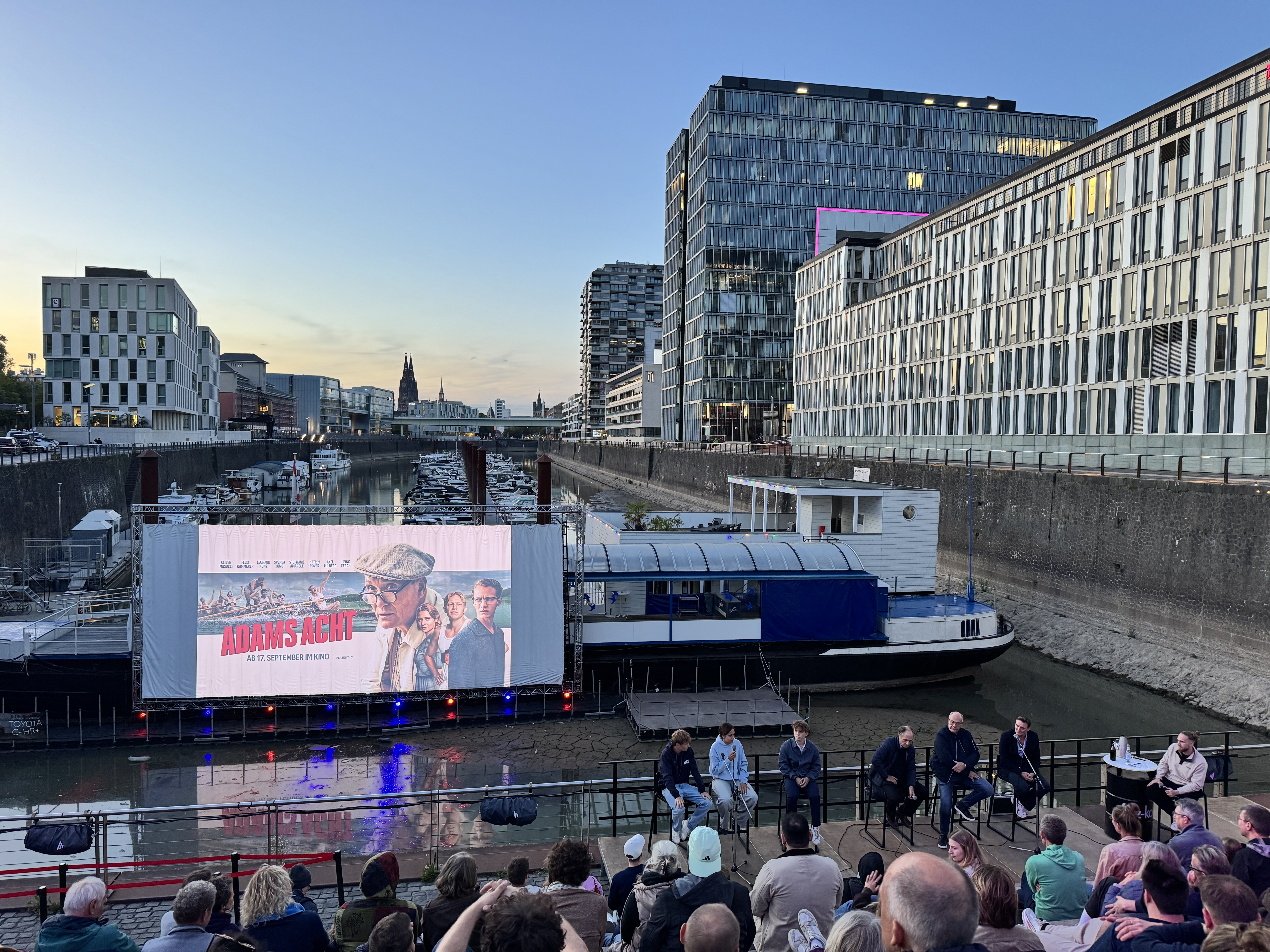 Das Bild zeigt die Talkrunde, die vor der Leinwand sitzt. Die Leinwand ist auf einem Ponton schwimmend vor einem Schiff montiert.