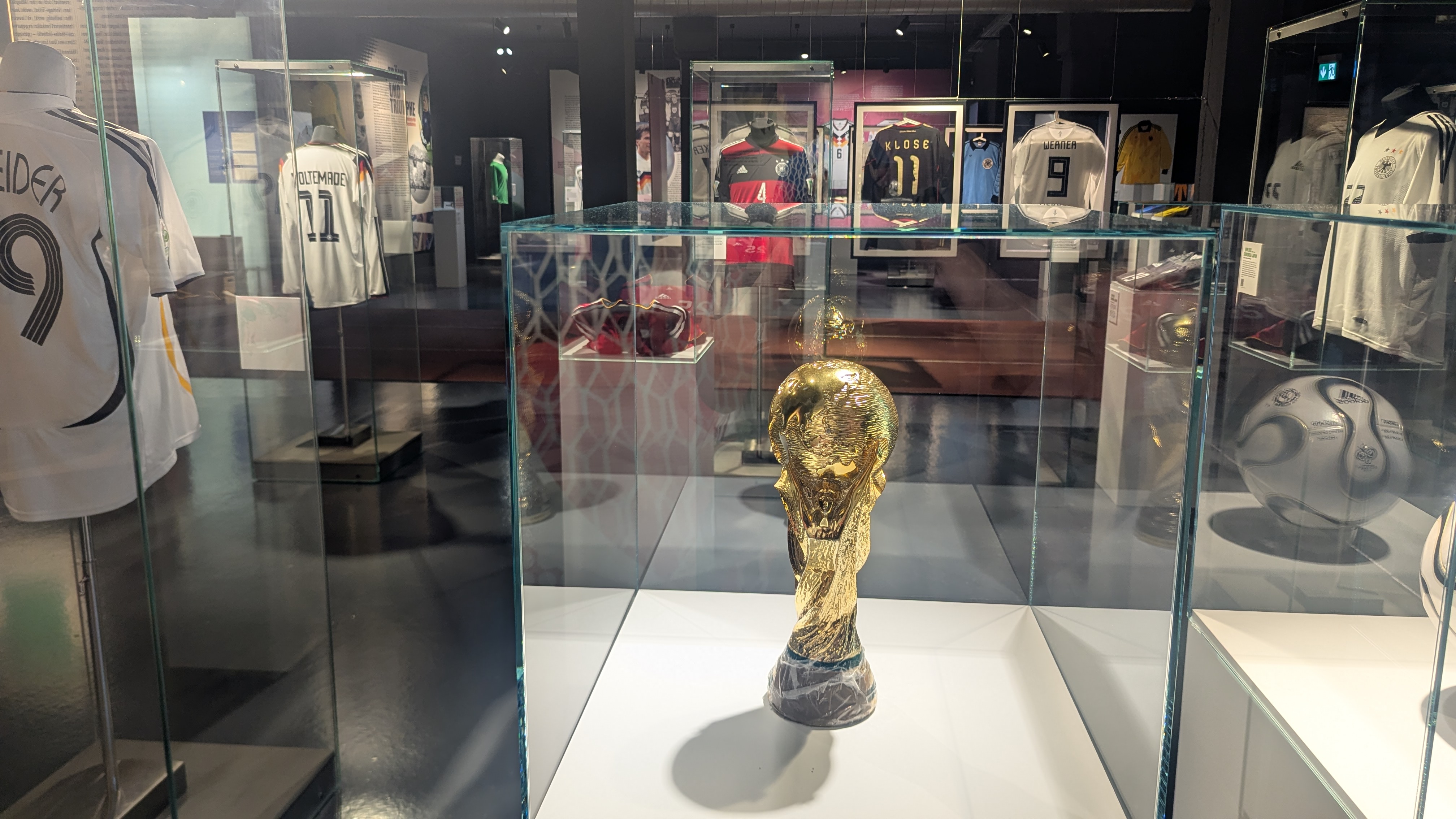 Ein Blick in die Ausstellung: Im Vordergrund steht eine NAchbildung des Weltpokals in einer Vitrine, dahinter sieht man verschieden Trikots in Bilderrahmen aufgehängt.