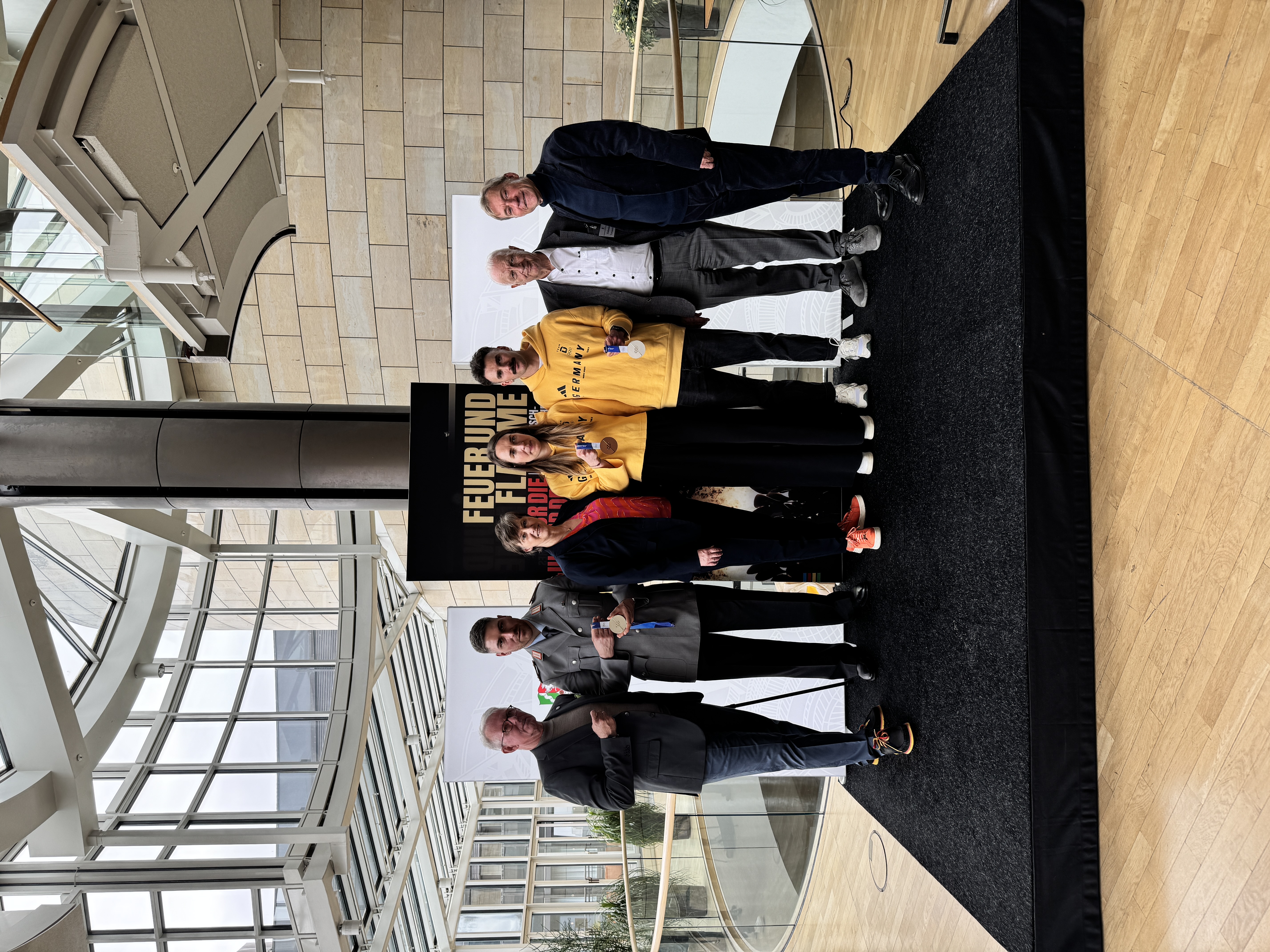 Es sind die sieben Personen nebeneinander auf der Bühne zu sehen. Die aktuellen Medaillengewinner:innen aus Milano Cortina zeigen ihre Medaillen.