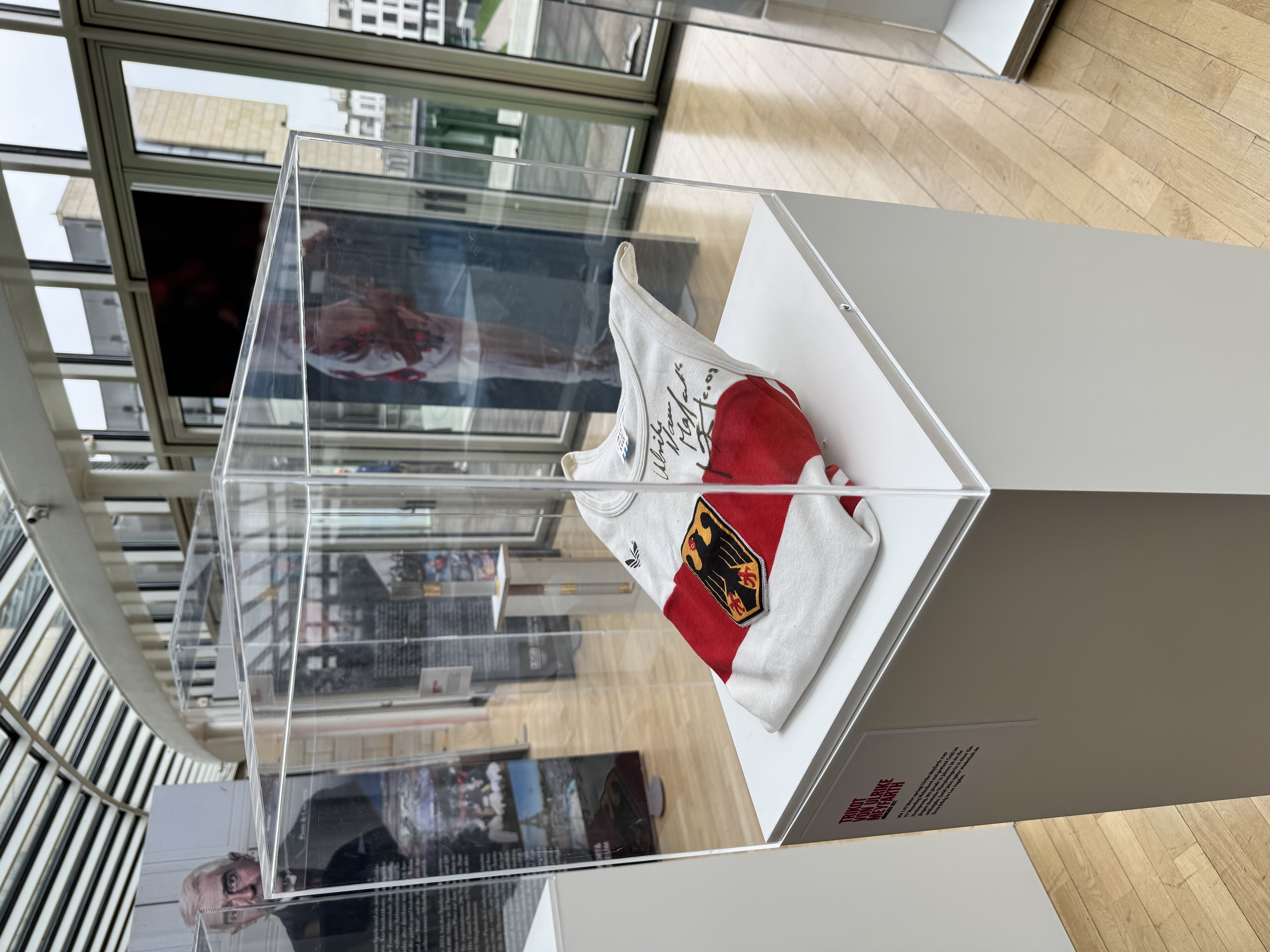 Trikot von Ulrike Meyfarth bein den Olympischen Spielen München 1972 in einer Sockelvitrine. Das Trikot ist weiß mit einem breiten roten Streifen und Bundesadler darauf.