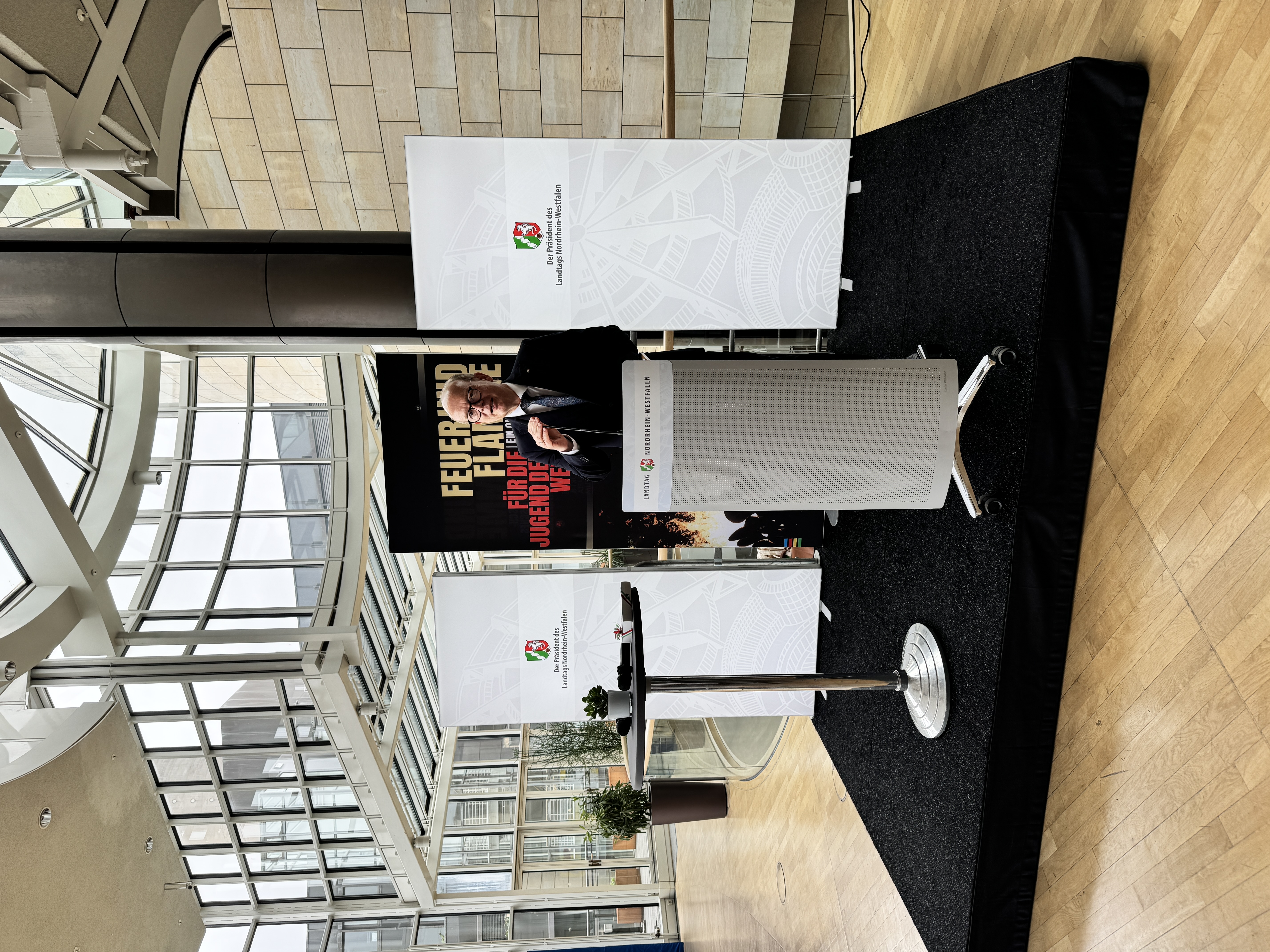 Blick auf die Bühne bei Eröffnung der Ausstellung. Dort zu sehen: Landtagspräsident André Kuper mit Mikrofon und Roll-Ups.