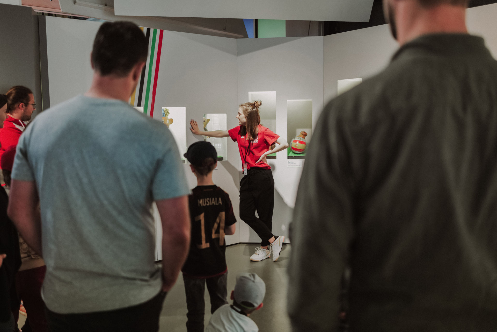 Es ist eine Führung in der Olympia Lounge. Eine Person mit roten Museumshirt erzählt etwas und Erwachsene und Kinder hören zu. Sie stehen mit dem Rücken zur Kamera.