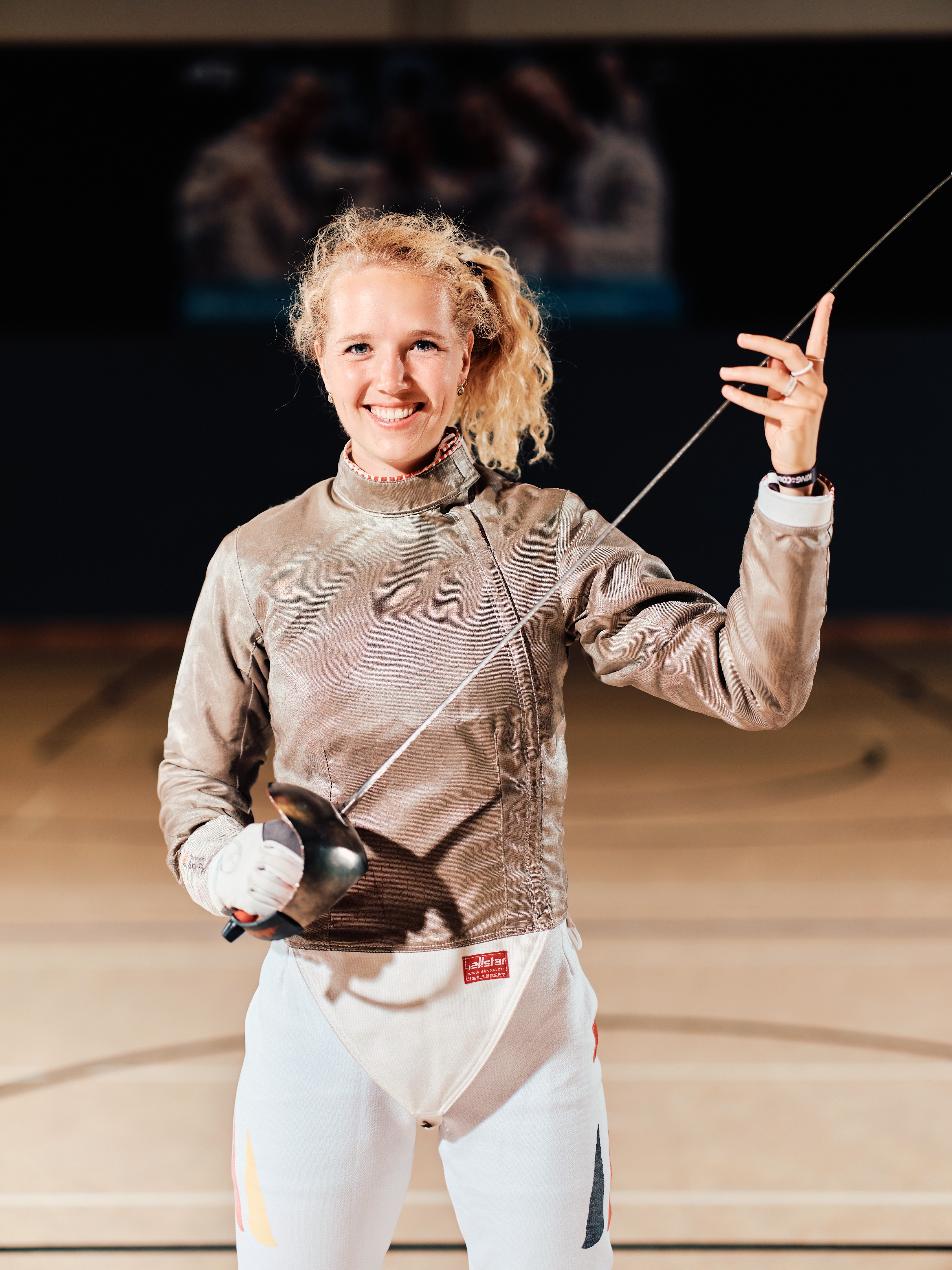 Auf dem Foto ist zentral Säbelfechterin Léa Krüger in ihrem Sportoutfit und mit Säbel zu sehen.