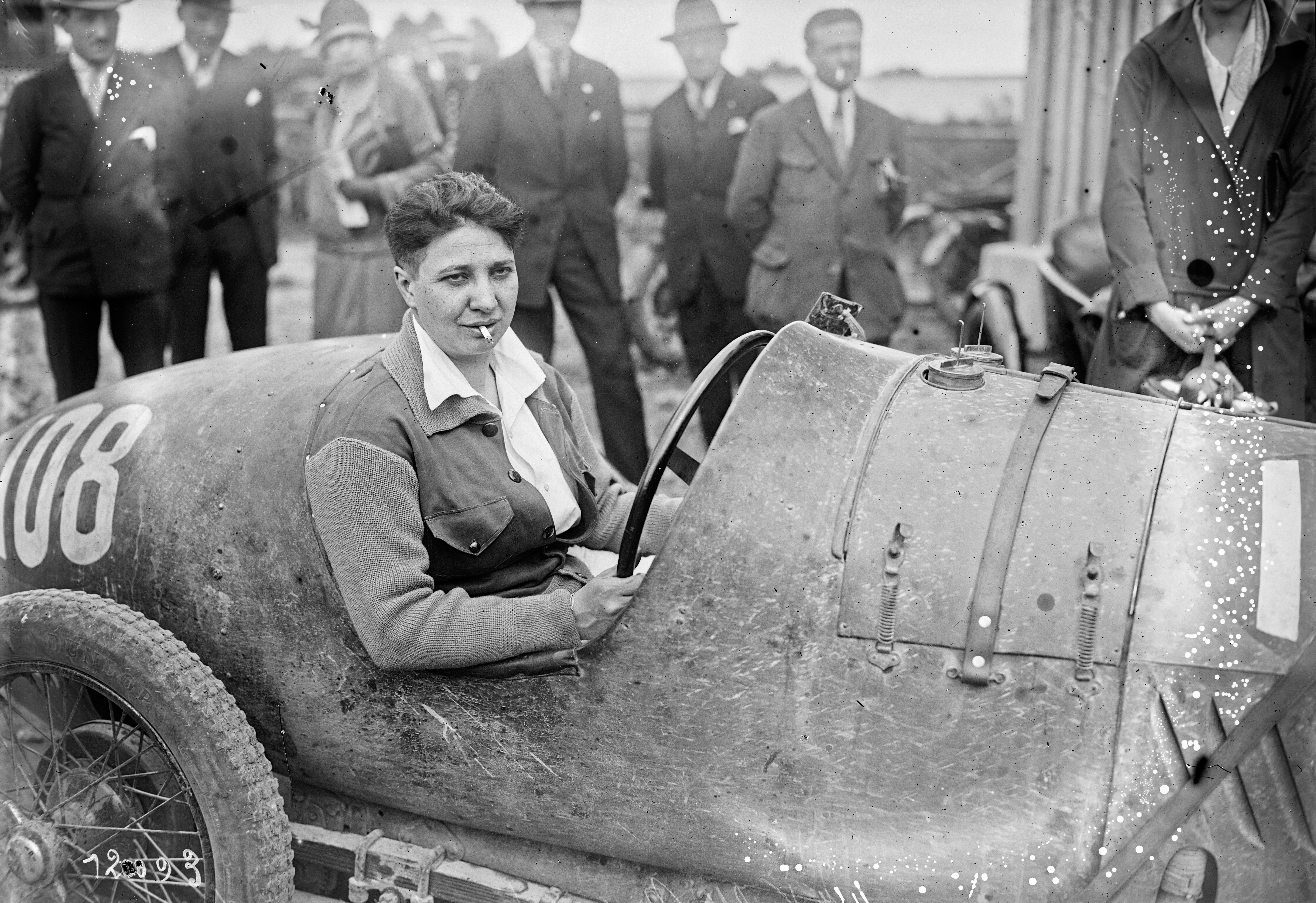 Das schwar-weiß Bild zeigt die Rennfahrerin Violette Morris 1927 in einem alten Rennwagen, umringt von mehreren Personen. Sie sitzt am Steuer und hat eine Zigarette im Mundwinkel.