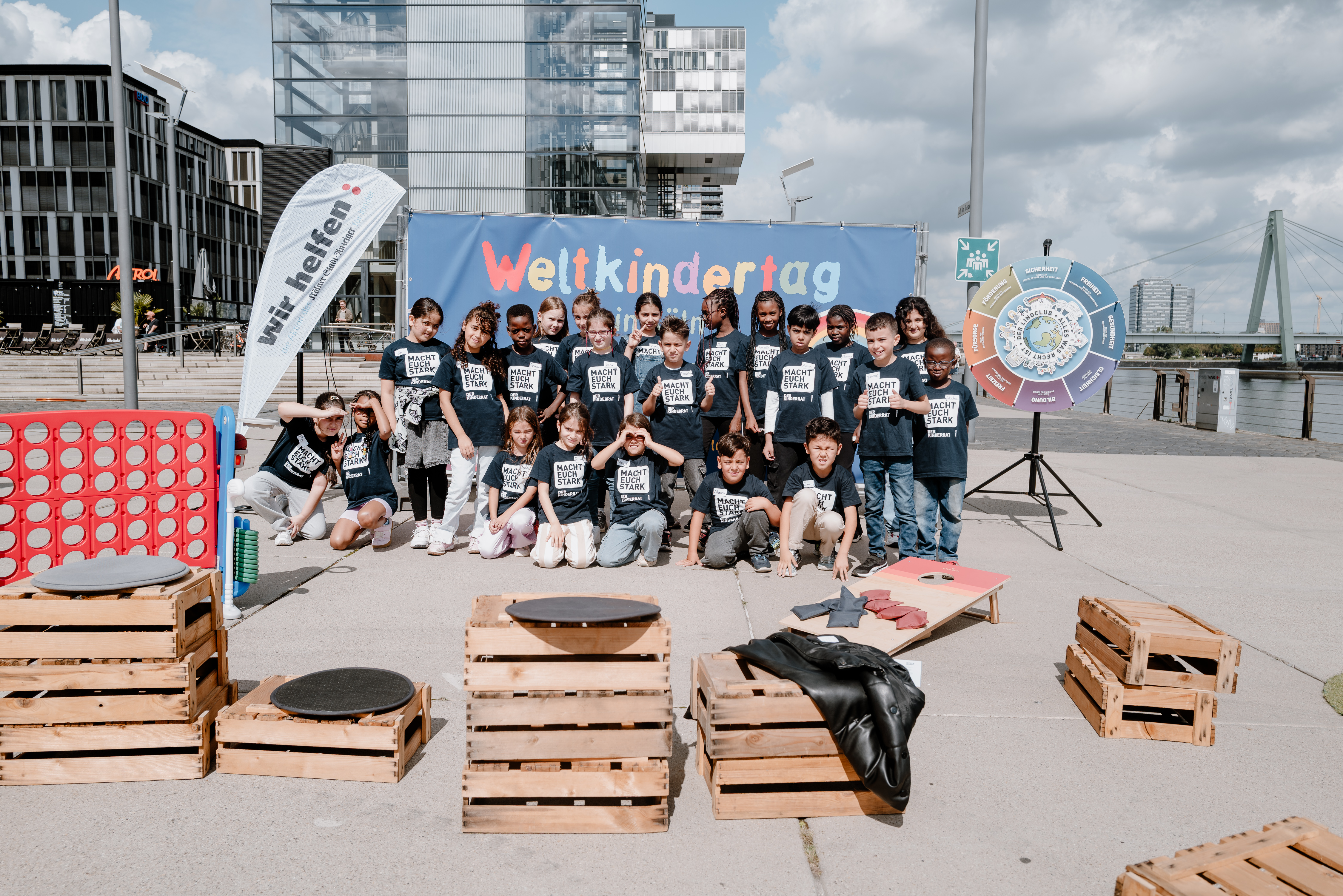 Zum Weltkindertag 2026 hat sich der Kinderrat für ein Foto aufgestellt. Die Kinder stehen und sitzen, sodass sie alle aufs Foto passen. Im Vordergrund sieht man verschiedene Spielstationen. Das Foto wurde im Rheinauhafen in Köln aufgenommen.