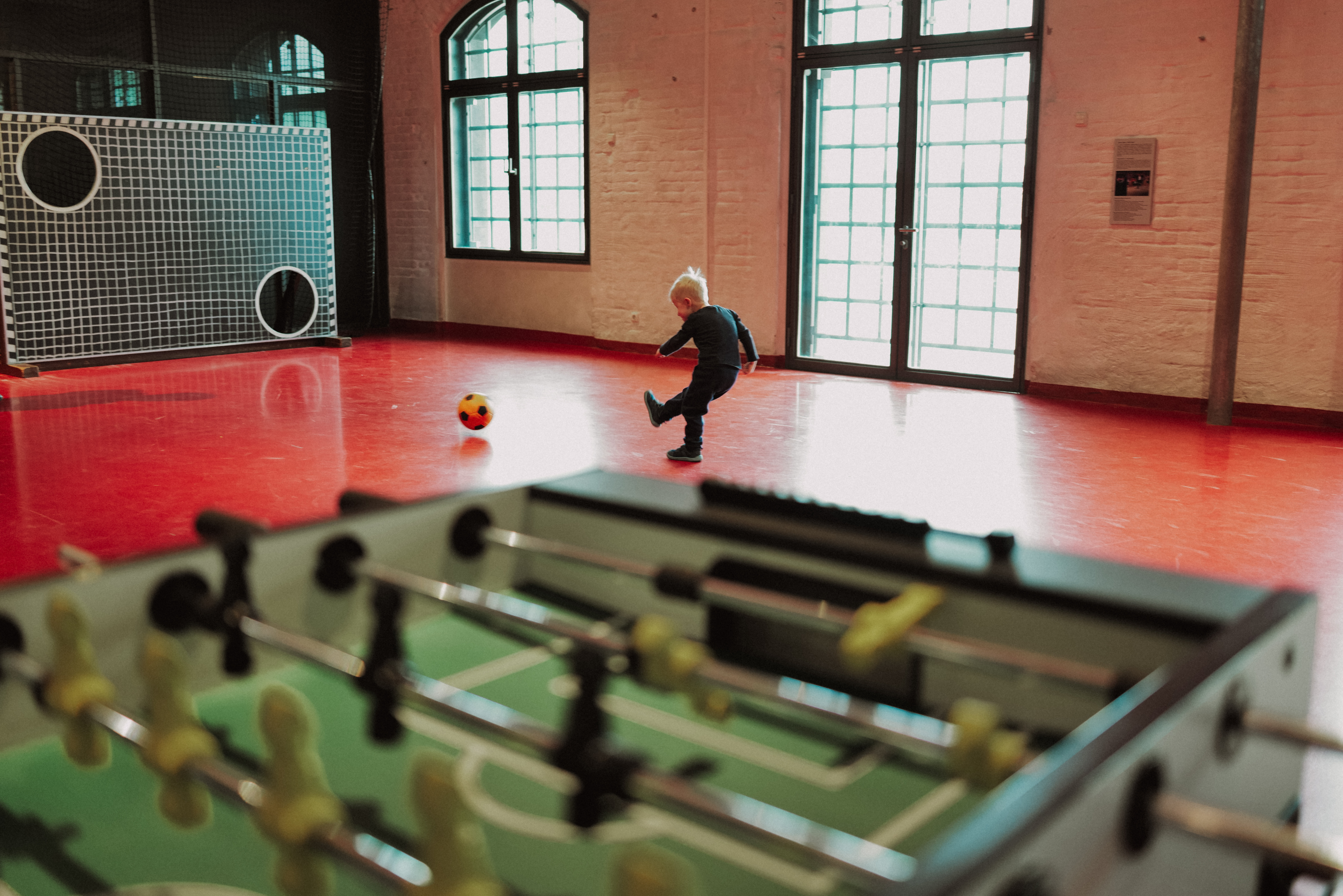 Im Vordergrund ist ein Tischkicker zu sehen. Im Hintergrund schießt ein kleiner Junge mit einem Ball auf eine Torwand.