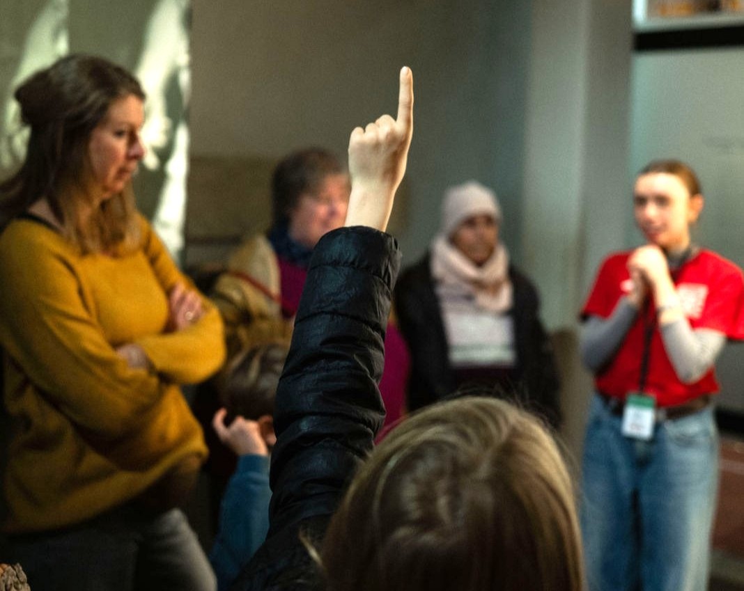 Es ist eine Gruppe bei einer Führung zu sehen. Ein Kind im Vordergrund ist von hinten zu sehen und hebt die Hand. Im Hintergrund ist ein Guide des Museums zu sehen.