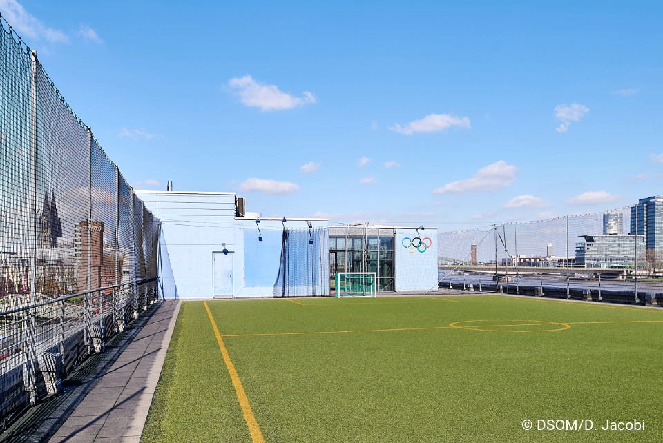 Das Foto wurde draußen gemacht. Es ist ein Fußballfeld auf dem Dach. Es steht ein kleines Tor auf dem Feld. Weiter hinten sind die Olympischen Ringe zusehen in den Farben Blau, Schwarz, Rot, Gelb und Grün. Neben dem Fußballspielfeld sind Netze gespannt. Der Kölner Dom ist links zu sehen und rechts der Rhein mit verschiedenen Häuseren.