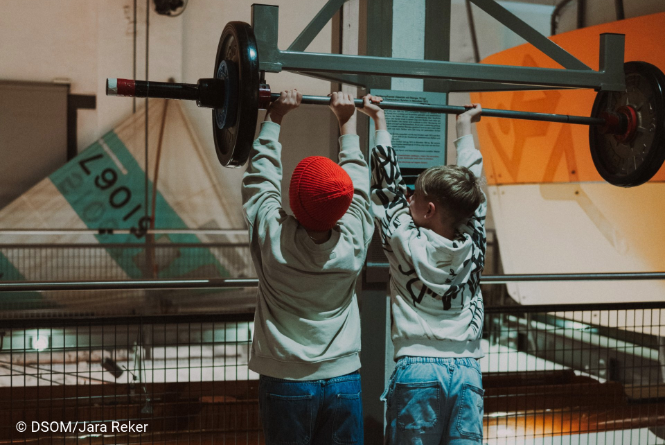 Es sind zwei Kinder zusehen. Beide Kinder sind an einer Langhantel. Die Langhantel hat an der Seite Gewicht. Sie versuchen die Langhantel hochzudrücken. Die Langhantel ist an der Wand befestigt, somit schwebt sie in der Luft. Im Hintergrund liegen zwei Segel.