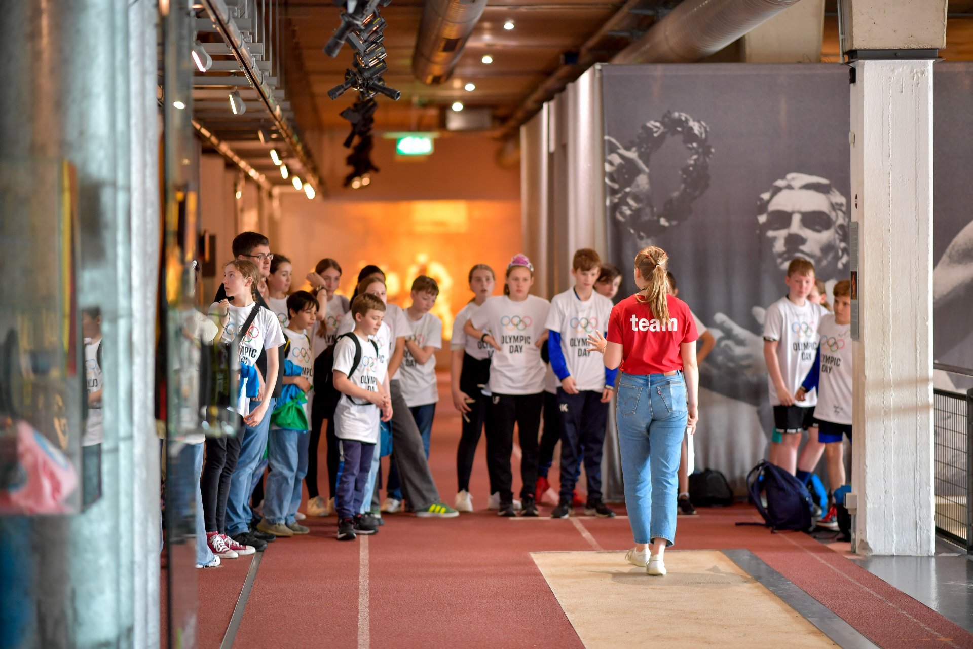 Das Bild worde in der Daueraustellung gemacht. In der Mitte steht eine Mitarbeiterin. Vor ihr steht eine Gruppe von Kinderen. Die Mitarbeiterin trägt ein rotes Shirt. Die Kinder haben ein weißes Shirt an. Passend zum Olympic Day. Sie hören der Mitarbeiterin aktiv zu. Sie stehen auf der Laufbahn.