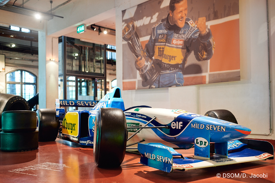Das Bild ist im Eingangsbereich entstanden. In der Mitte steht ein Formel 1 Wagen. Um den Wagen herrum, stehen weitere Reifen. An der Wand hängt eine Leinwand. Auf der Leinwand ist Michael Schumacher mit einen Pokal zu sehen . Im Hintergrund sind große Fester und ein Fahrstuhl.
