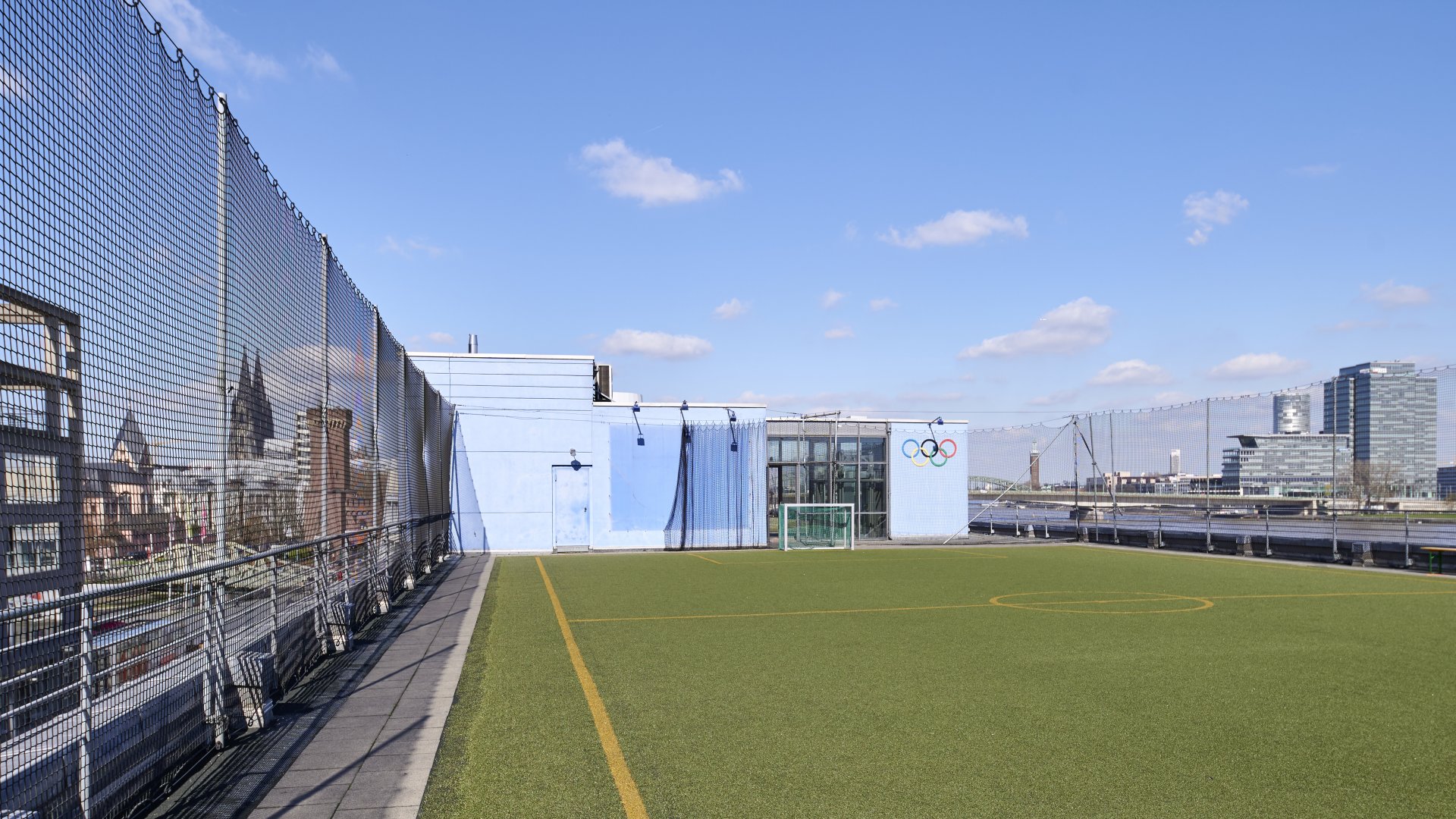 Das Foto wurde draußen gemacht. Es ist ein Fußballfeld auf dem Dach. Es steht ein kleines Tor auf dem Feld. Weiter hinten sind die Olympischen Ringe zusehen in den Farben Blau, Schwarz, Rot, Gelb und Grün. Neben dem Fußballspielfeld sind Netze gespannt. Der Kölner Dom ist links zu sehen und rechts der Rhein mit verschiedenen Häuseren.