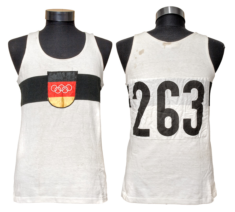 Weißes Leichtathletiktrikot mit schwarzem Bruststreifen sowie Wappen mit olympischen Ringen auf Schwarz, Rot, Gold