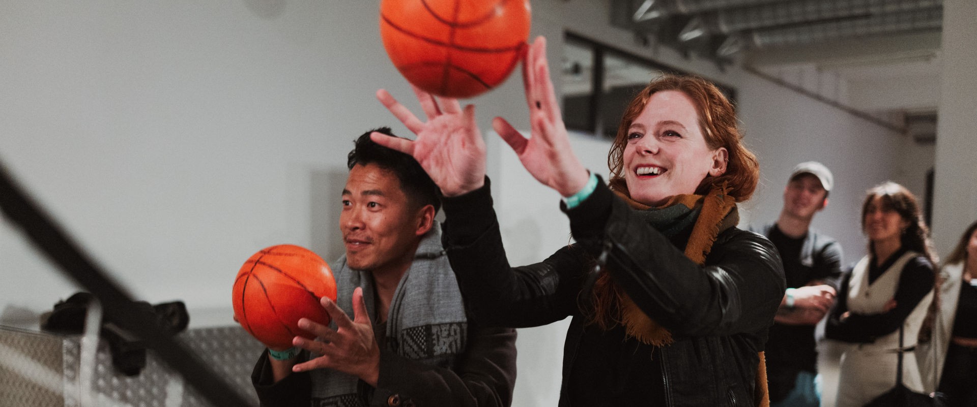 Es spielen zwei Personen drinnen Basketball. Vorne steht eine Frau und daneben ein Mann. Beide werfen den Basketball in Richtung Korb. Hinter den beiden, stehen viele Menschen im Hintergrund. Der Raum ist groß und bieten immer noch genügt Platz.