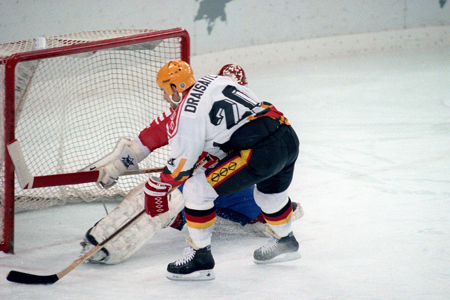 In dem Bild sind zwei Eishockeyspieler, die gegeneinander spielen. Der Torwart ist Kanadier und der Feldspieler ist Deutscher. Einer ist Torwart und der andere Spieler ist Feldspieler. Der Feldspieler hat ein Deutschlandtrikot an. Er versucht, ein Tor zu schießen Der Torwart steht im Tor und hält den Puck.