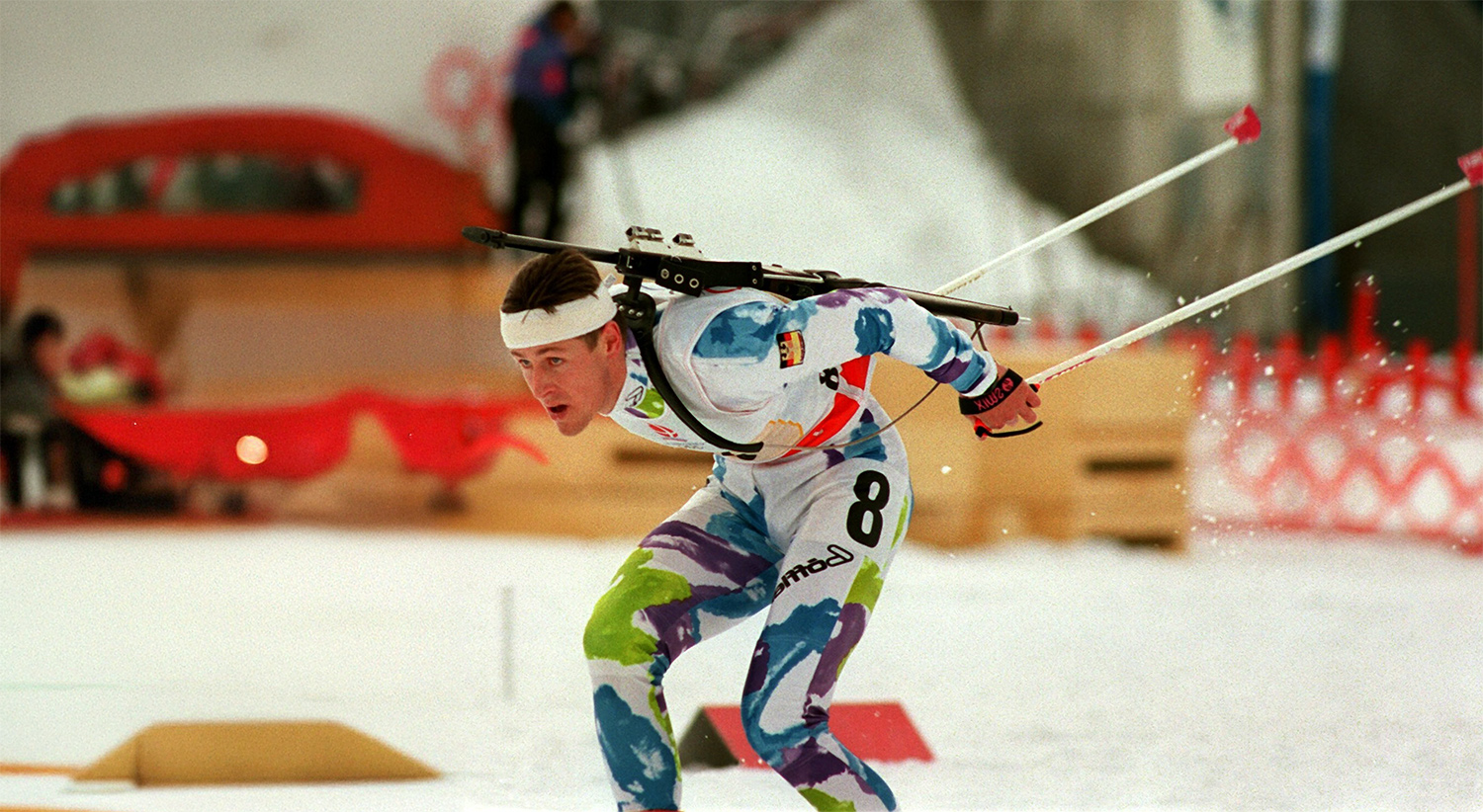 In dem Bild ist ein Biathlet. Er fährt gerade auf seinen Skiern und hat auf dem Rücken sein Gewehr In seinen Händen hält er seine Langlaufstöcke. Sein Anzug hat verschiedene Farben. Seine Startnummer ist die Nummer acht. Im Hintergrund sind ein paar Menschen und ein Zaun. Auf dem Foto ist recht viel Schnee zu sehen.