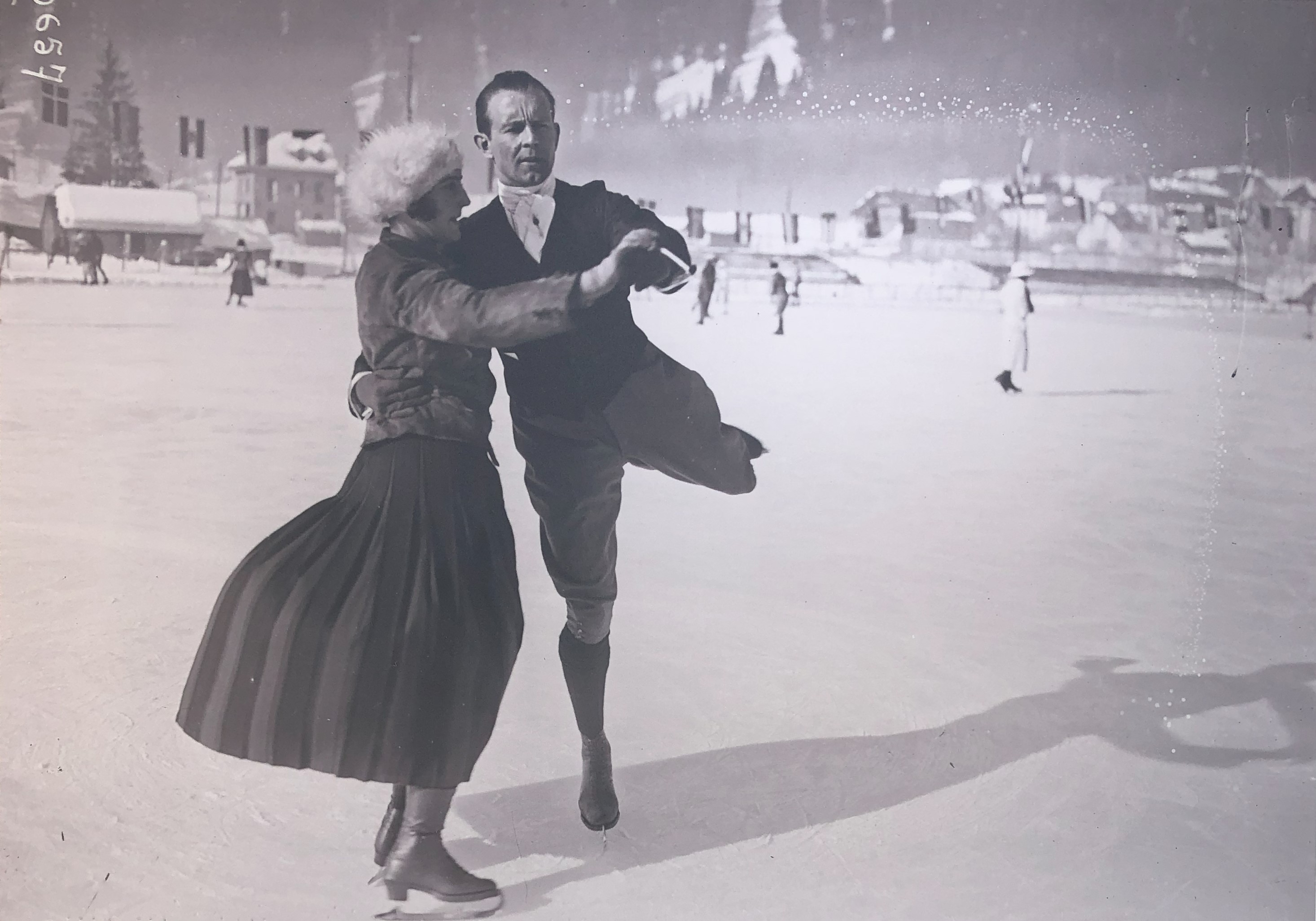 Das Bild ist Schwarz-Weiß. Es sind eine Frau und ein Mann im Vordergrund. Die beiden Personen fahren zusammen Schlittschuh. Die Frau und der Mann halten sich an der Hand. Der Mann hat ein Bein in der Luft und das andere Bein ist auf der Eisfläche. Im Hintergrund fahren auch andere Paare zusammen.
