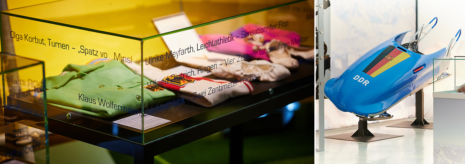 On the left side of the picture is a glass display case with various exhibits. The exhibits are from the 1972 Olympic Games in Munich. On the right is a blue bobsled from the GDR. In front is the flag of the GDR, and under the flag is the abbreviation DDR.