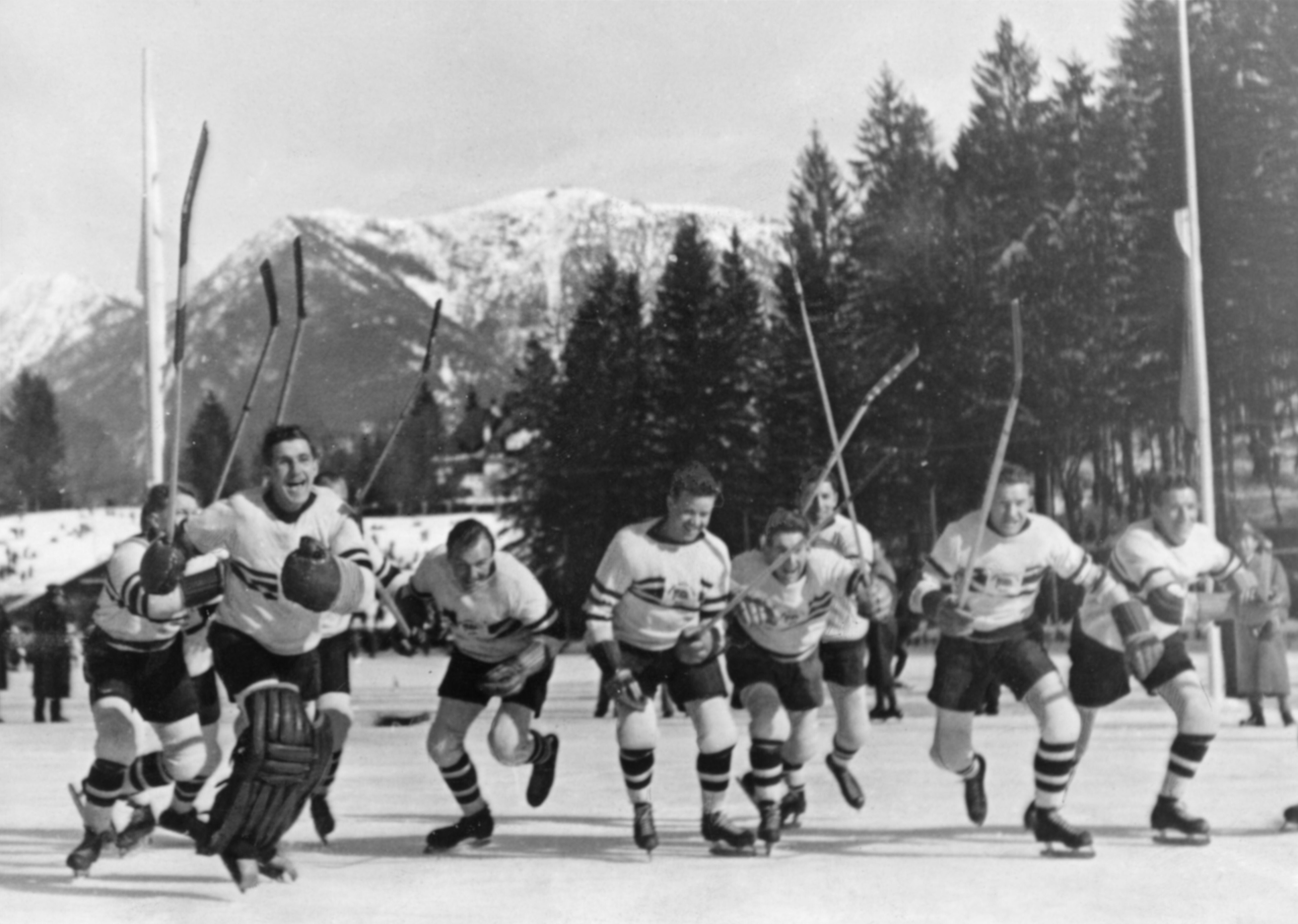 Das Bild ist Schwarz-Weiß. Es ist eine Eishockeymannschaft zu sehen Die Mannschaft steht auf dem Eis und freut sich. Die Spieler laufen in Richtung des Fotografen Im Hintergrund sind Bäume und Berge zu sehen.