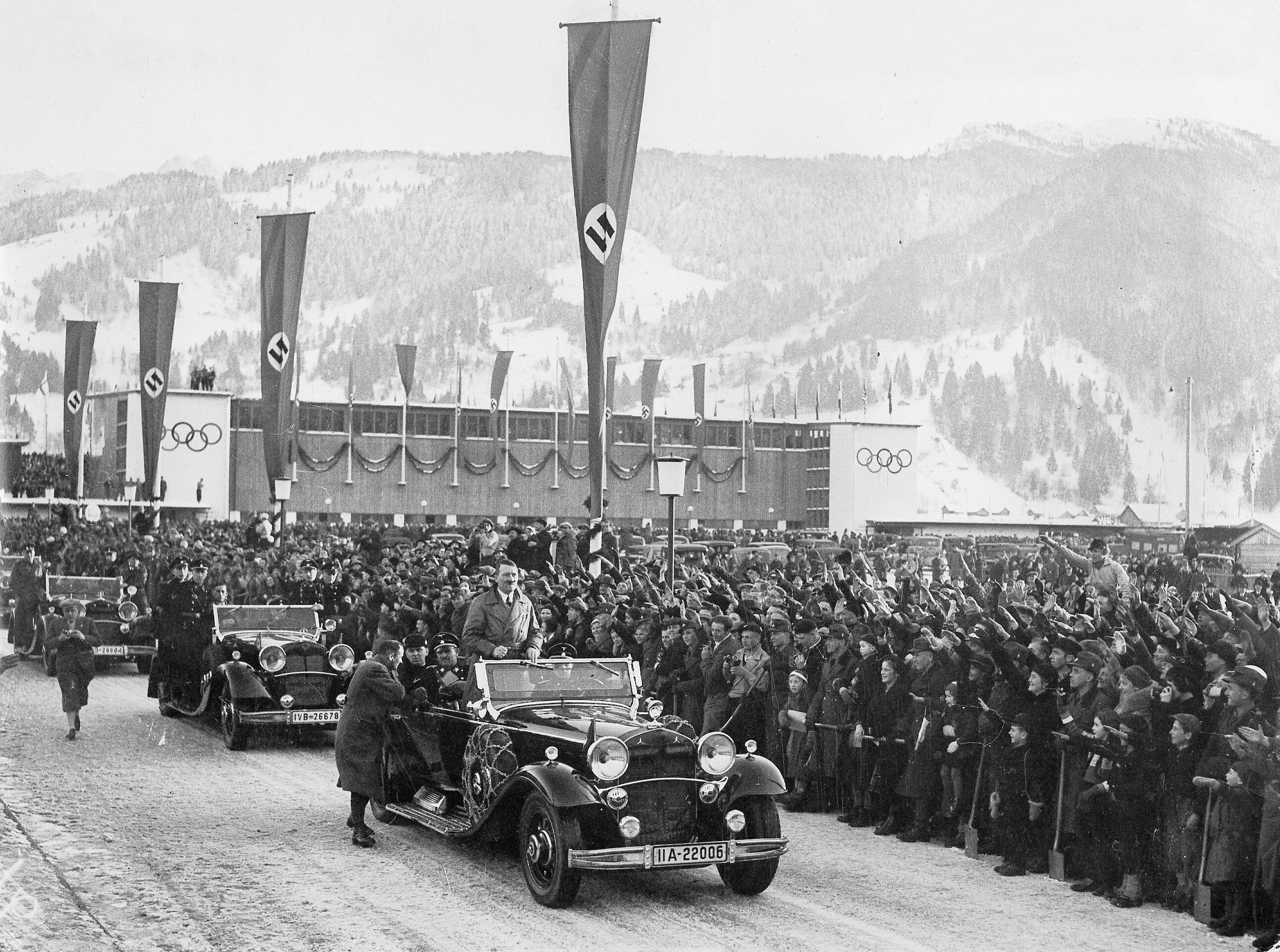 Das Bild ist Schwarz-Weiß. Es sind mehrere Autos zu sehen Im ersten Auto steht Adolf Hitler. Neben den Autos stehen ganz viele Menschen und jubeln ihm zu. Es sind auch verschiedene Fahnen aufgehängt worden, mit einem Hakenkreuz. Im Hintergrund ist ein Gebäude mit den Olympischen Ringen. Weiterhin sind auch Berge zu sehen.