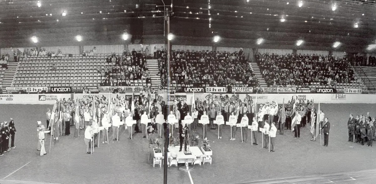 Das Bild ist Schwarz-Weiß. Es ist die Eröffnung von den Paralympics 1976. Die Tribünen sind nur auf der rechten Seite gefüllt. Die linke Seite ist leer. Auf dem Rasen stehen ein paar Athleten. Vor ihnen stehen kleine Kinder mit Schildern.