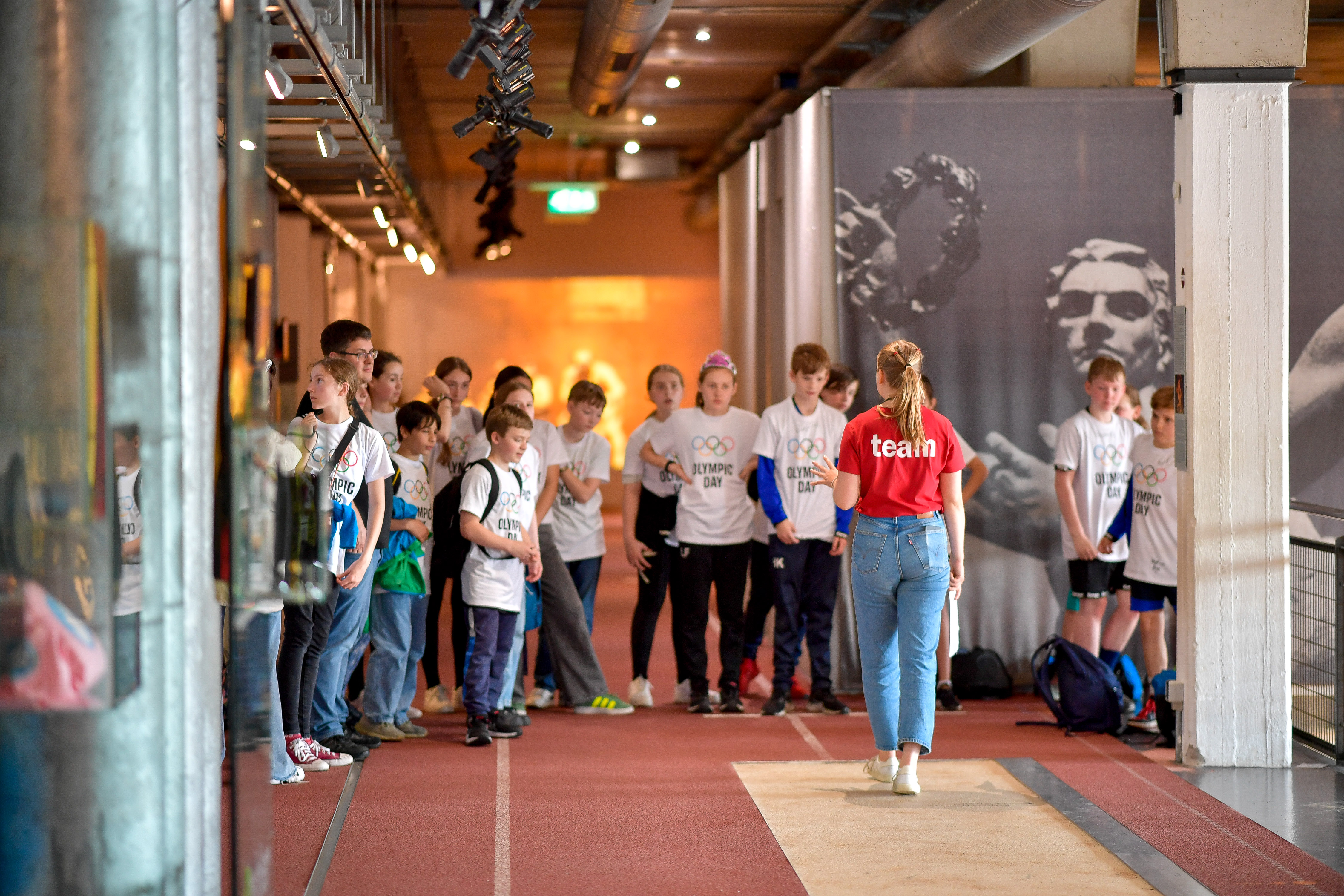 Das Bild wurde in der Dauerausstellung gemacht. In der Mitte steht eine Mitarbeiterin. Vor ihr steht eine Gruppe von Kindern. Die Mitarbeiterin trägt ein rotes Shirt. Die Kinder haben ein weißes Shirt an. Passend zum Olympic Day. Sie hören der Mitarbeiterin aktiv zu. Sie stehen auf der Laufbahn.