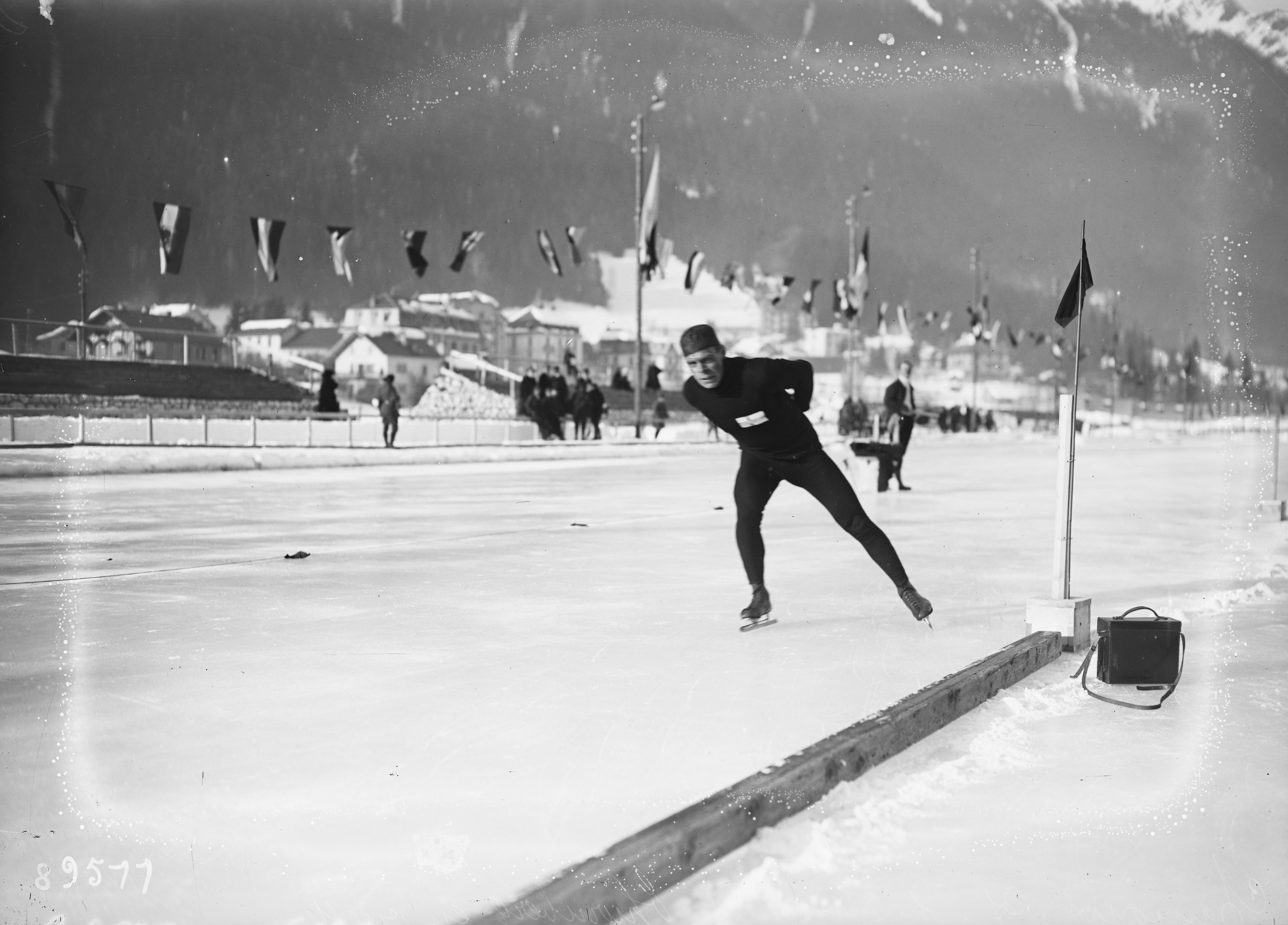 Das Foto ist Schwarz-Weiß. Es ist ein Eisschnellläufer zu sehen Er fährt gerade auf der Eisbahn. Neben der Bahn sind verschiedene Fahnen. Im Hintergrund ist ein Berg und verschiedene Menschen.